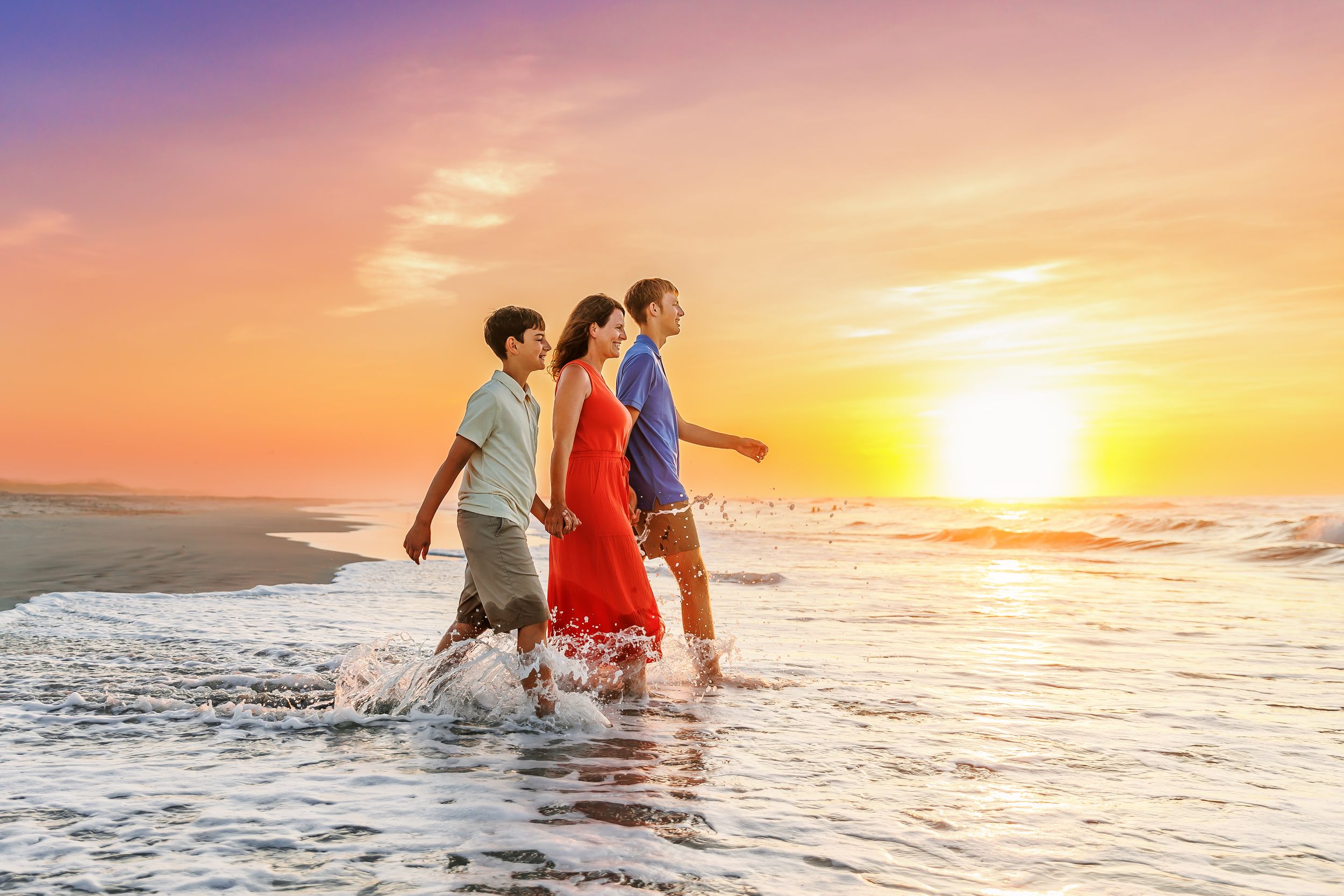 Three people walking together along the beach shoreline at sunset with orange sky and waves splashing at their feet.