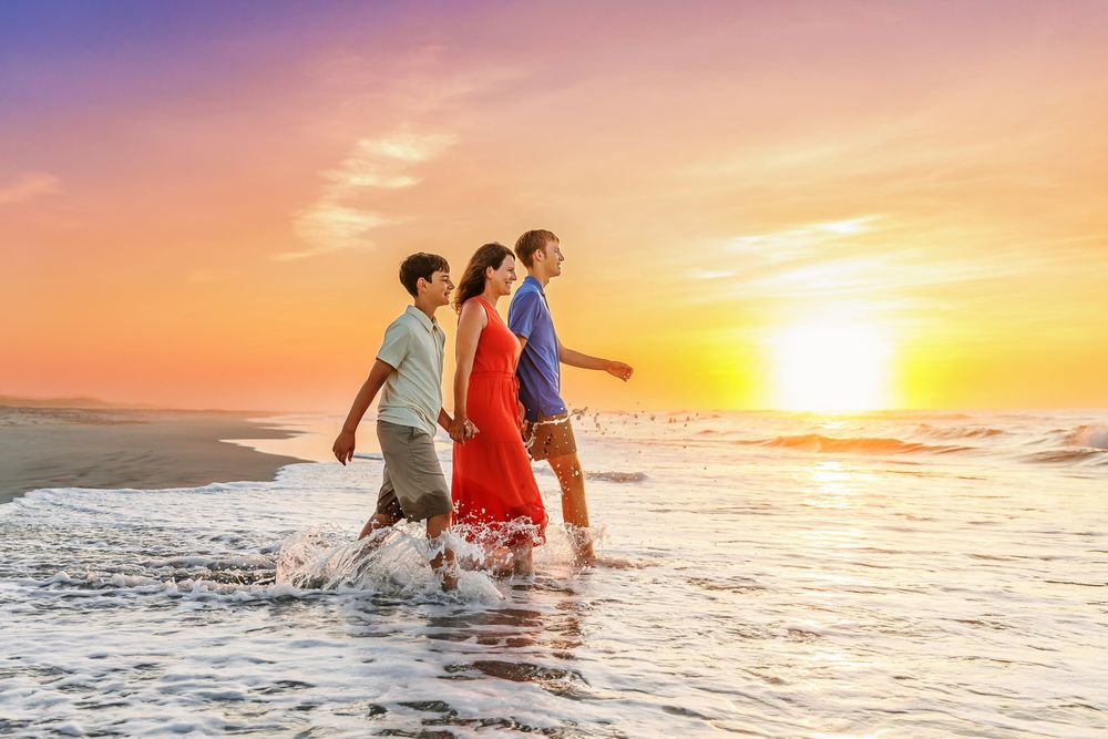 Three people walking together along the beach shoreline at sunset with orange sky and waves splashing at their feet.