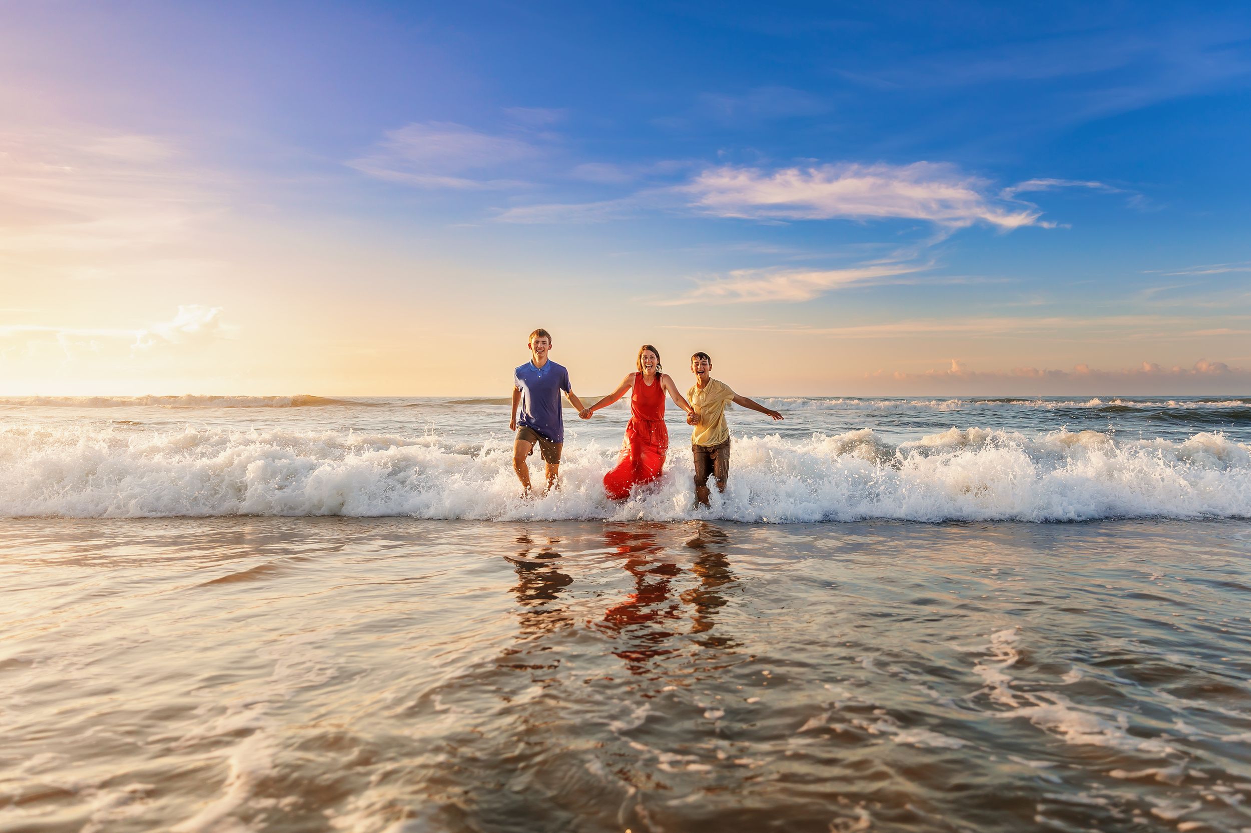 Three people holding hands while running through ocean waves at sunset on a beautiful beach.