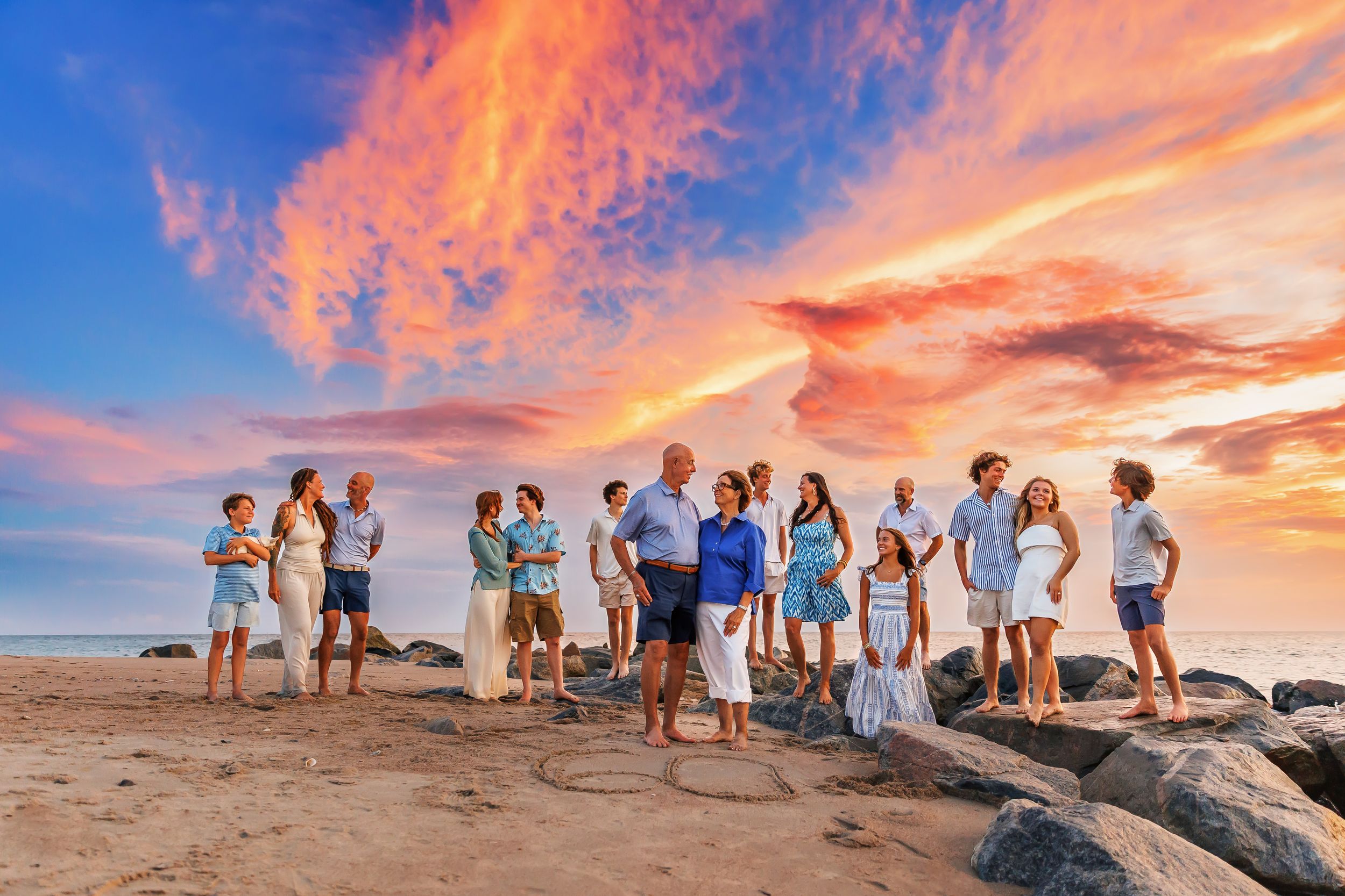 A group of people standing on a beach at sunset with dramatic orange and pink clouds in the sky.