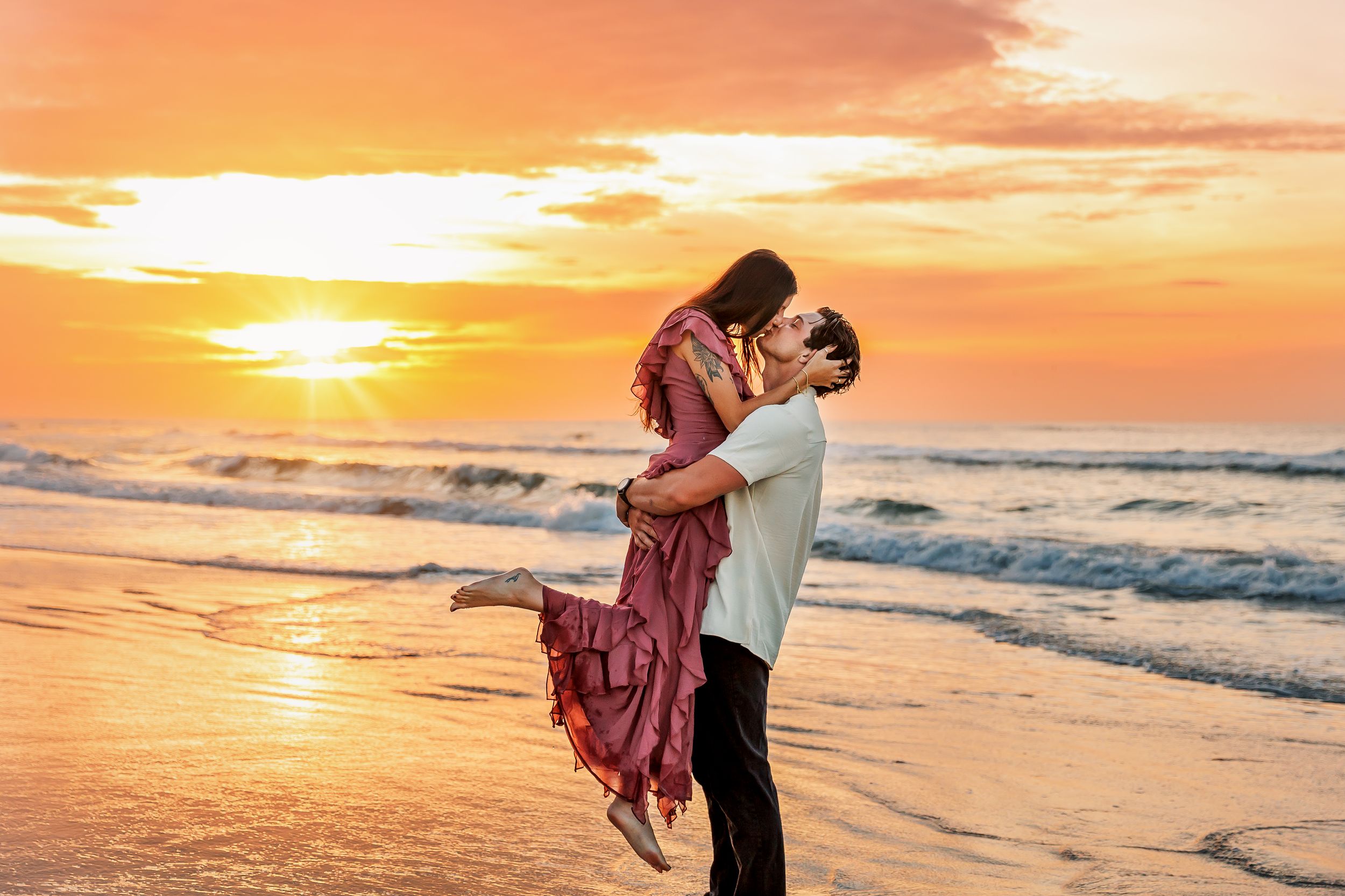 Two silhouettes embrace on a beach at sunset as golden light reflects off gentle waves rolling onto shore.