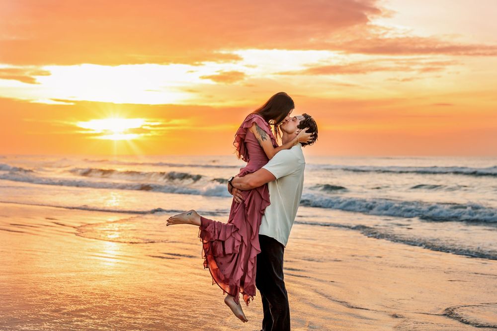 Two silhouettes embrace on a beach at sunset as golden light reflects off gentle waves rolling onto shore.