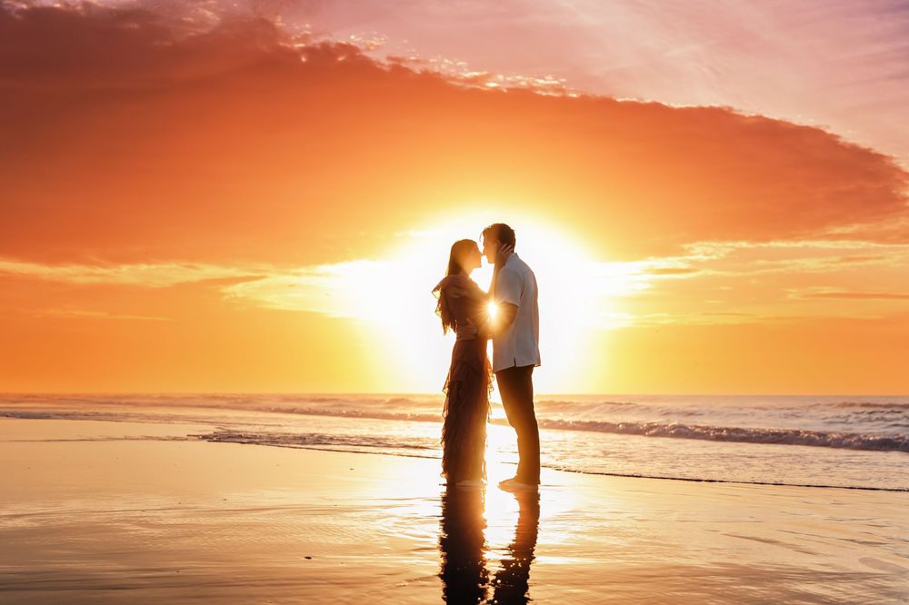 Silhouetted couple embraces on beach at sunset with orange sky and ocean waves in background.