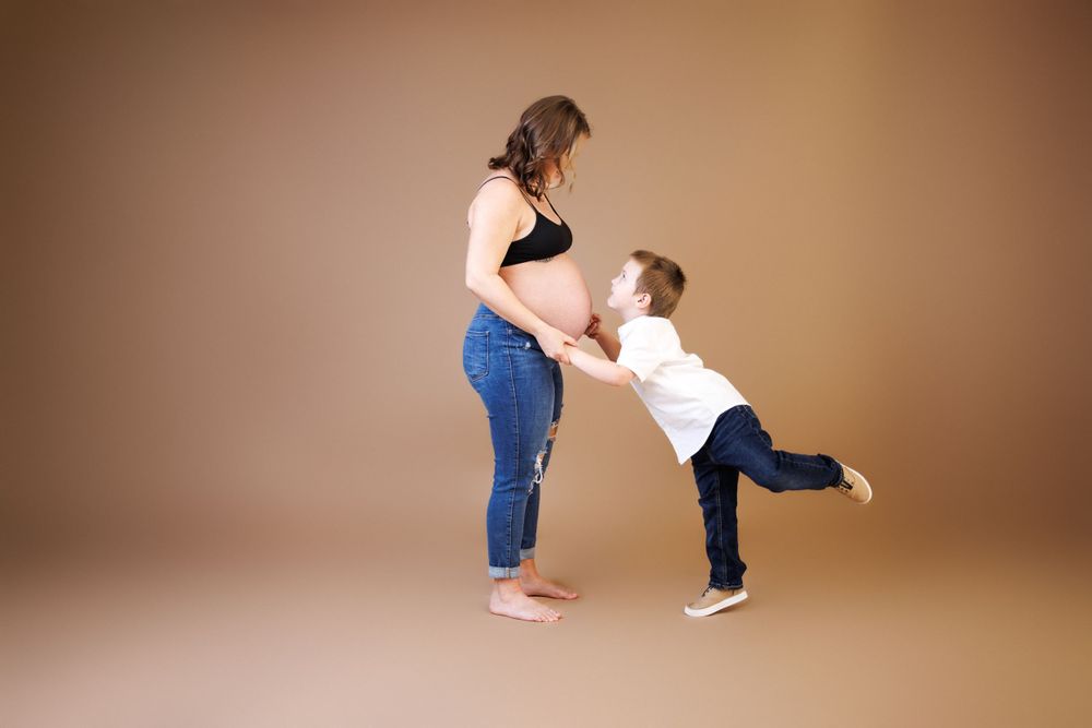 A pregnant woman in jeans and a black top stands with her child who leans forward playfully against a brown backdrop.