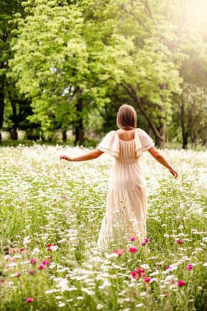 Woman in flowy beige dress standing among wildflowers and daisies in a sunlit meadow on a summer day.