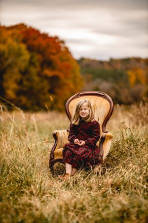 Child sitting in vintage chair placed in autumn field with fall colors.
