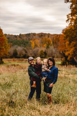 Family of four standing together in fall field with colorful autumn foliage.
