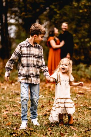 Two children walking hand in hand through autumn leaves with adults in background.