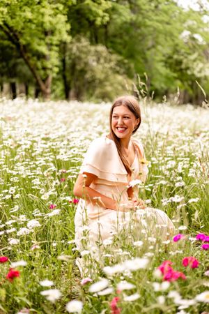 Person in peach dress sitting joyfully among blooming wildflowers and daisies in a garden setting.