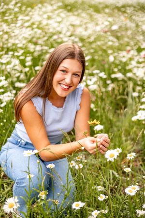 Someone in casual denim outfit kneeling to pick daisies in a blooming summer meadow.