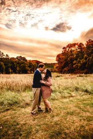 Couple embracing in golden field during dramatic sunset with fall foliage.