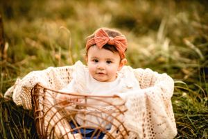 Baby wearing bow headband sitting in woven basket outdoors.