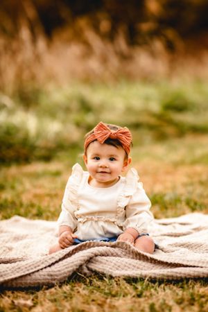 Baby with orange bow headband sitting on blanket outdoors.