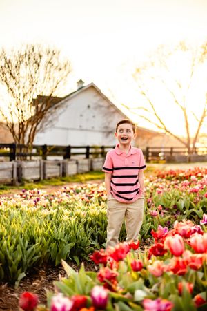 Young person standing among colorful tulip fields at sunset with white barn in background.