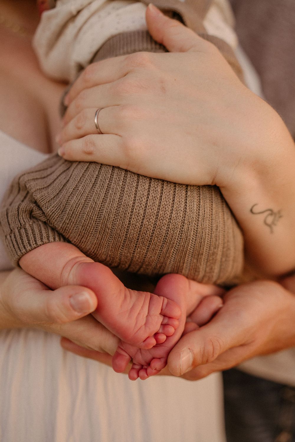 Close-up of tiny baby feet held in parent's hands with delicate ring and tattoo detail.