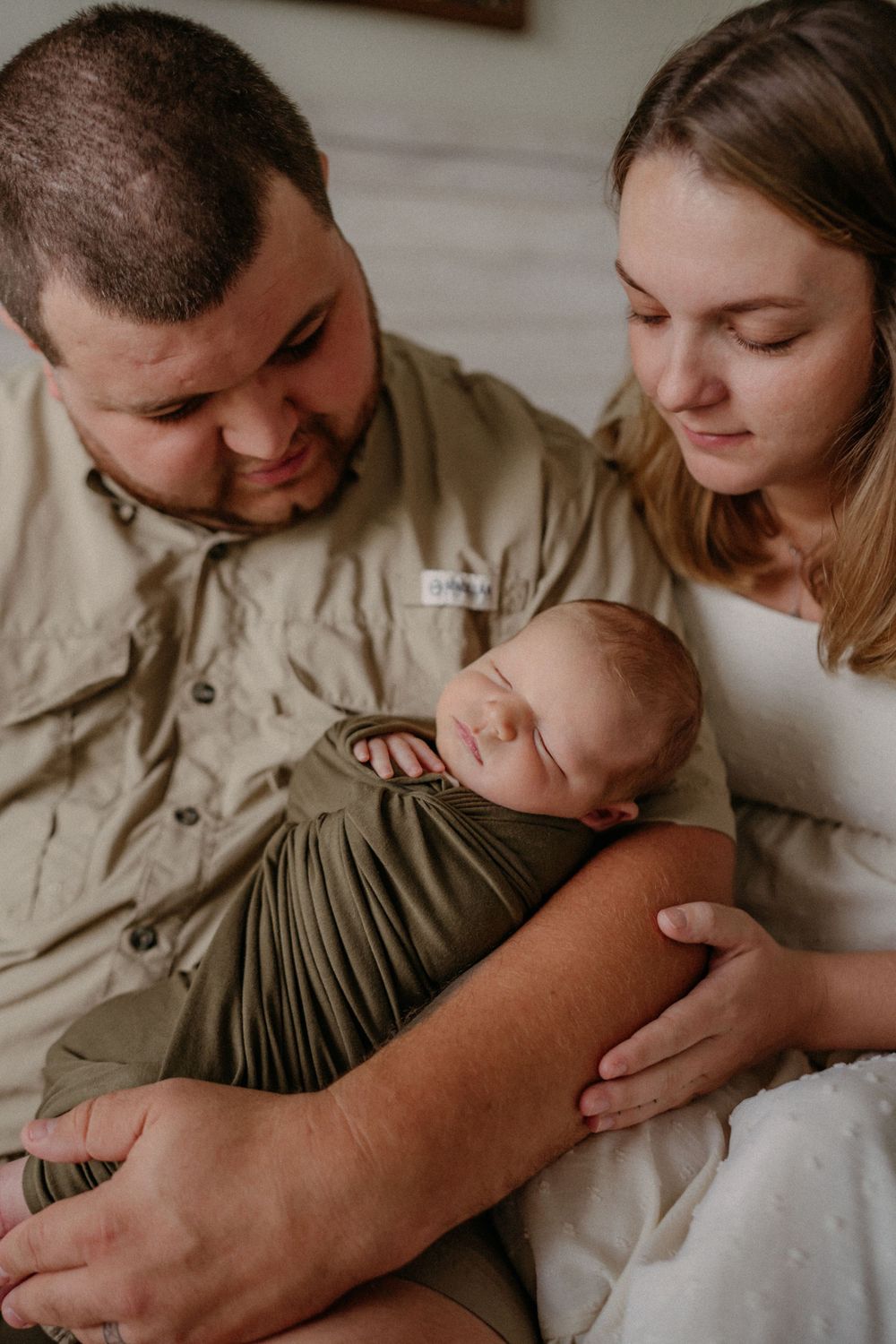 A sleeping newborn swaddled in olive green fabric is cradled gently by parents in a tender indoor family moment.