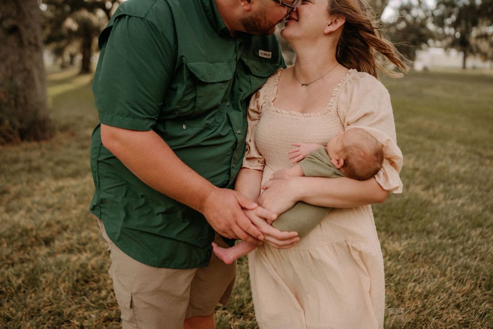 Couple embracing in an outdoor field while holding and nursing a newborn baby at sunset.