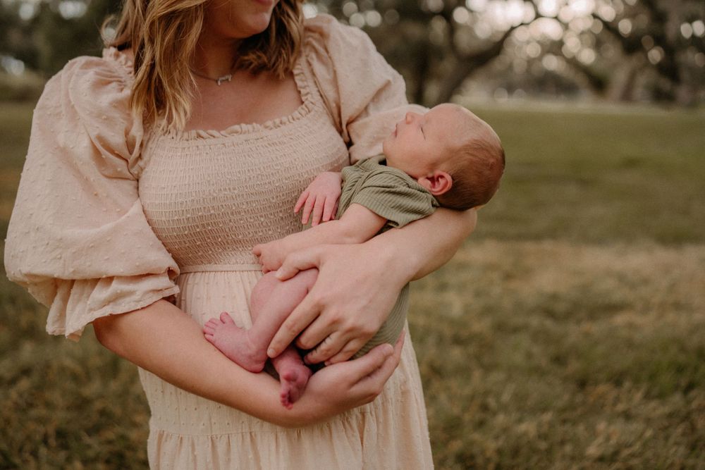 A sleeping infant being cradled in arms while wearing a light green outfit against a soft outdoor background.