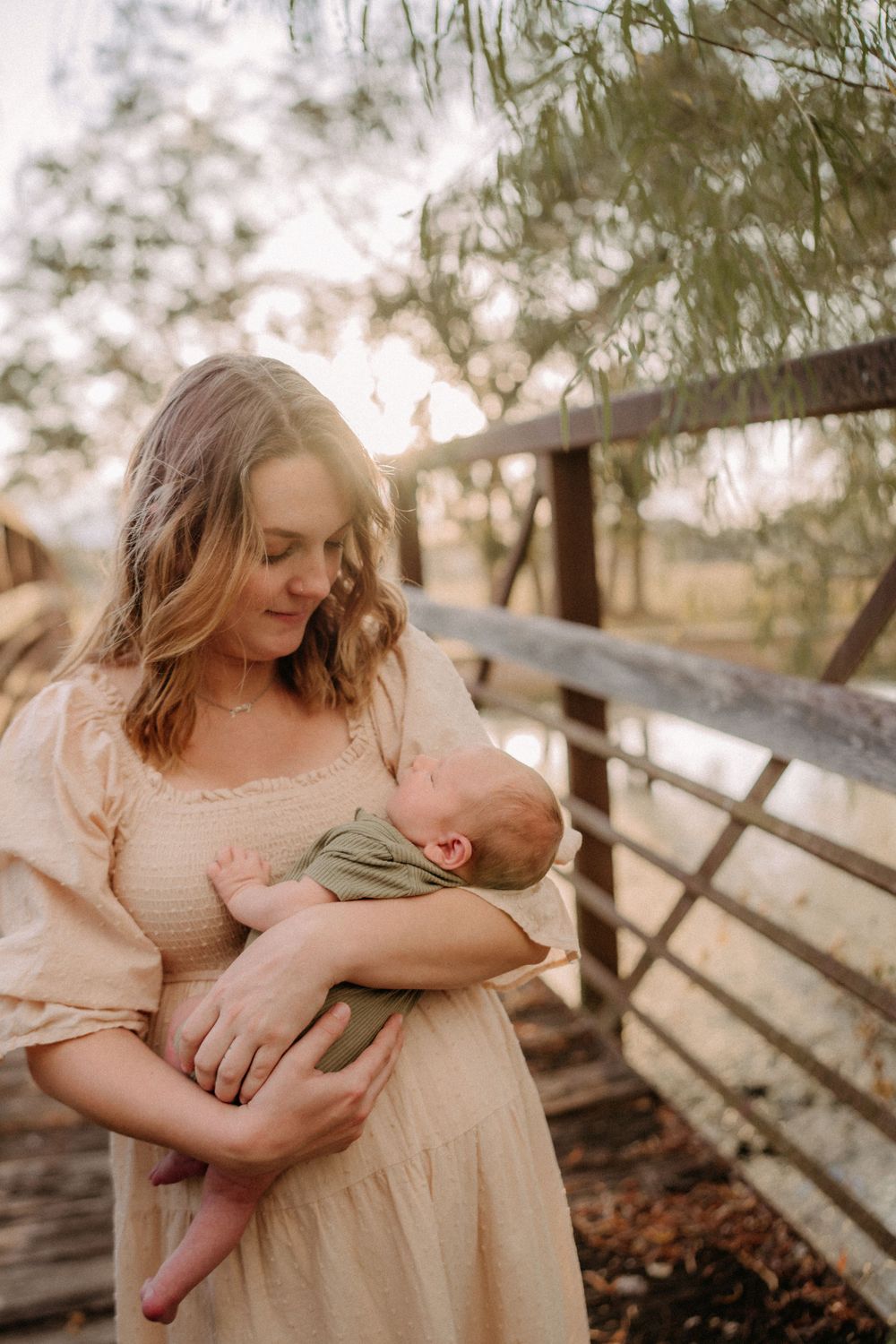 Parent in cream dress holding sleeping newborn near rustic wooden fence at sunset.