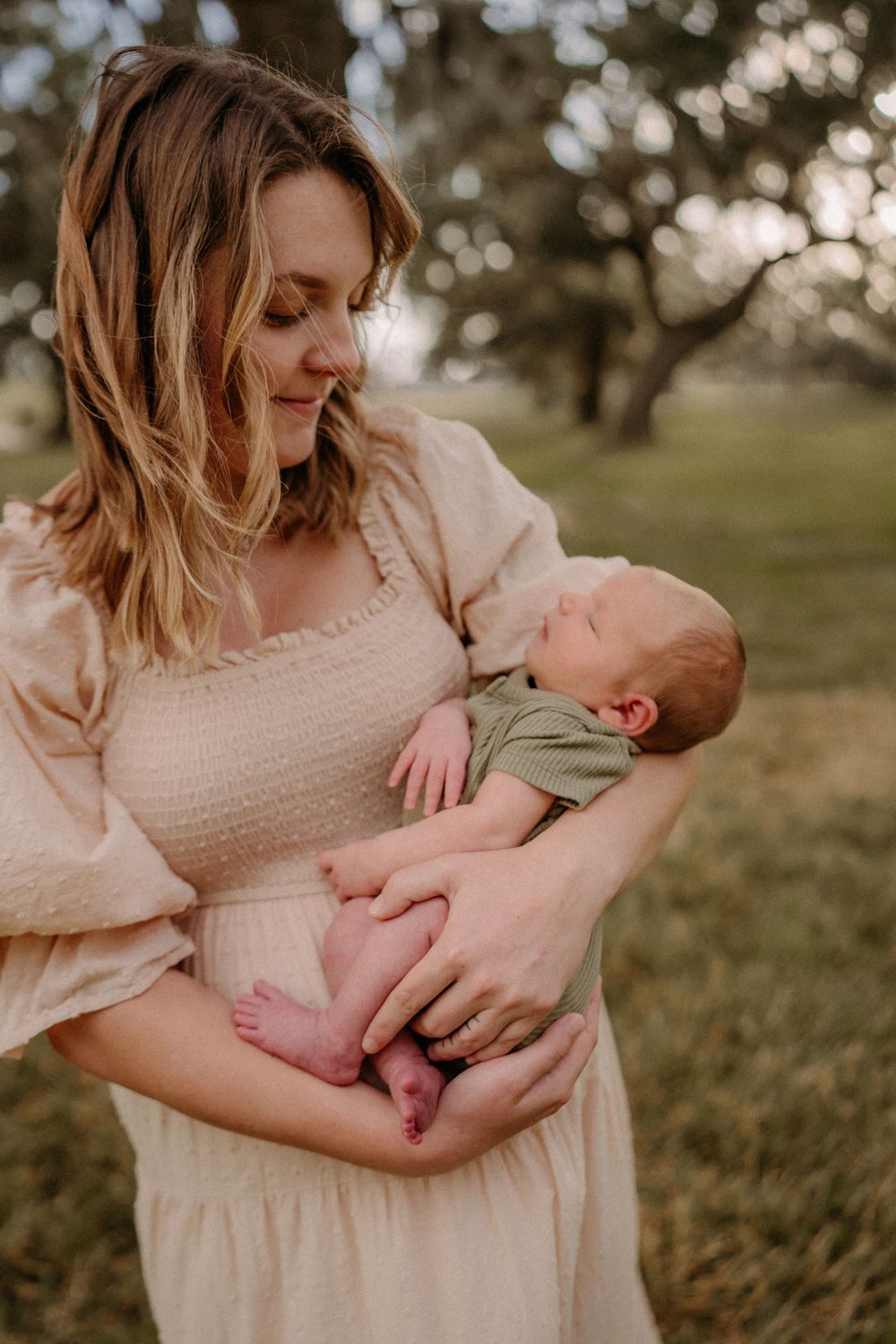 A tender moment between parent and newborn baby outdoors at sunset in a cream-colored dress.