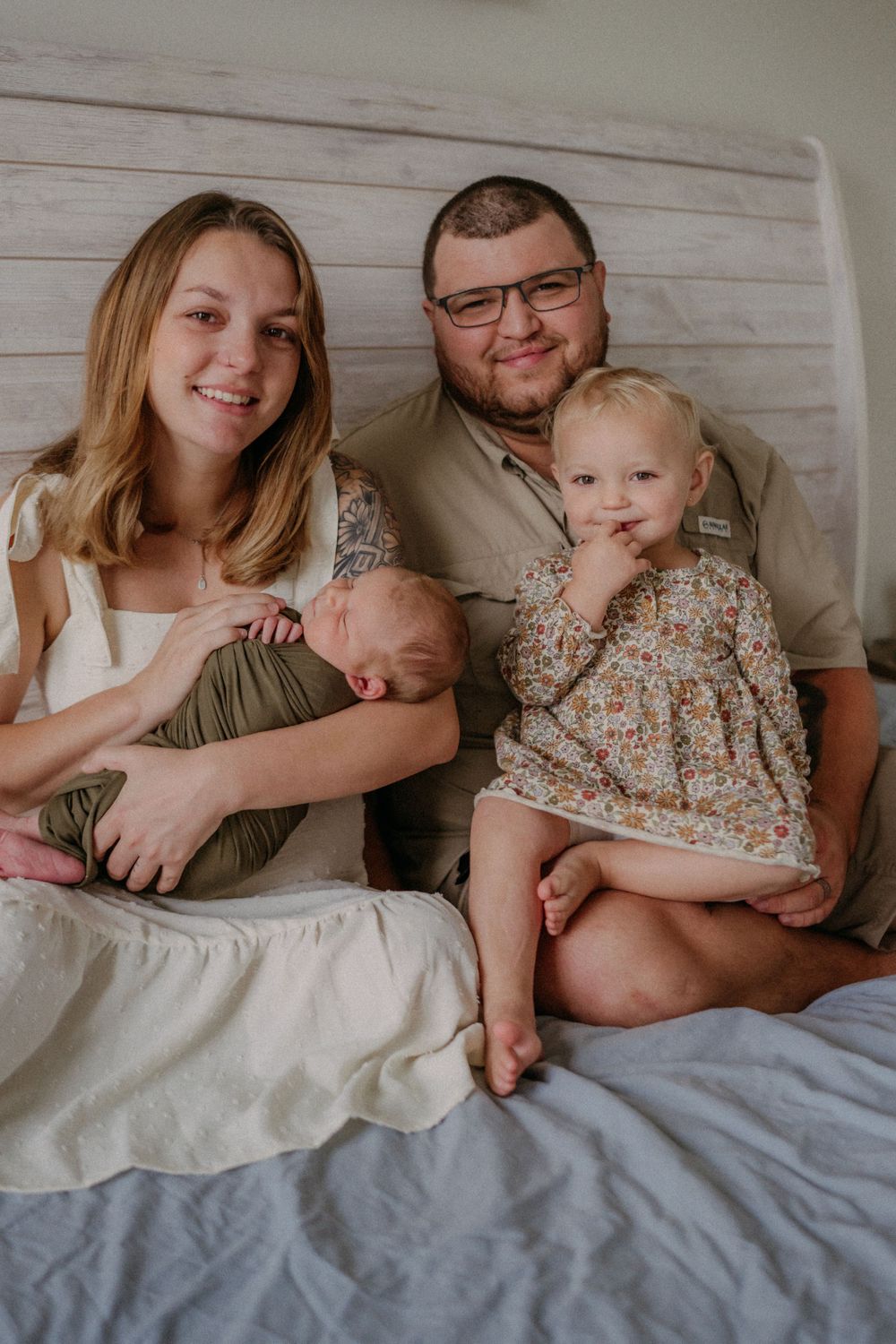 A family with two small children poses together on a bed with white shiplap wall background.