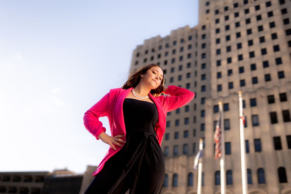 Figure in bright pink jacket poses dramatically against urban building backdrop.