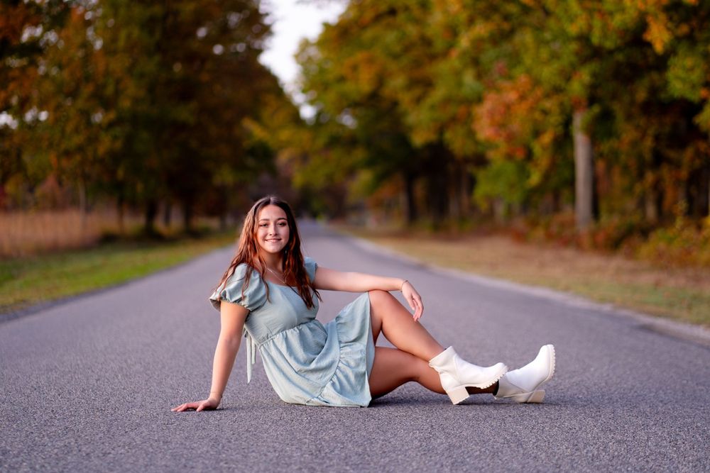 Person in casual dress poses on empty road with autumn trees in background.