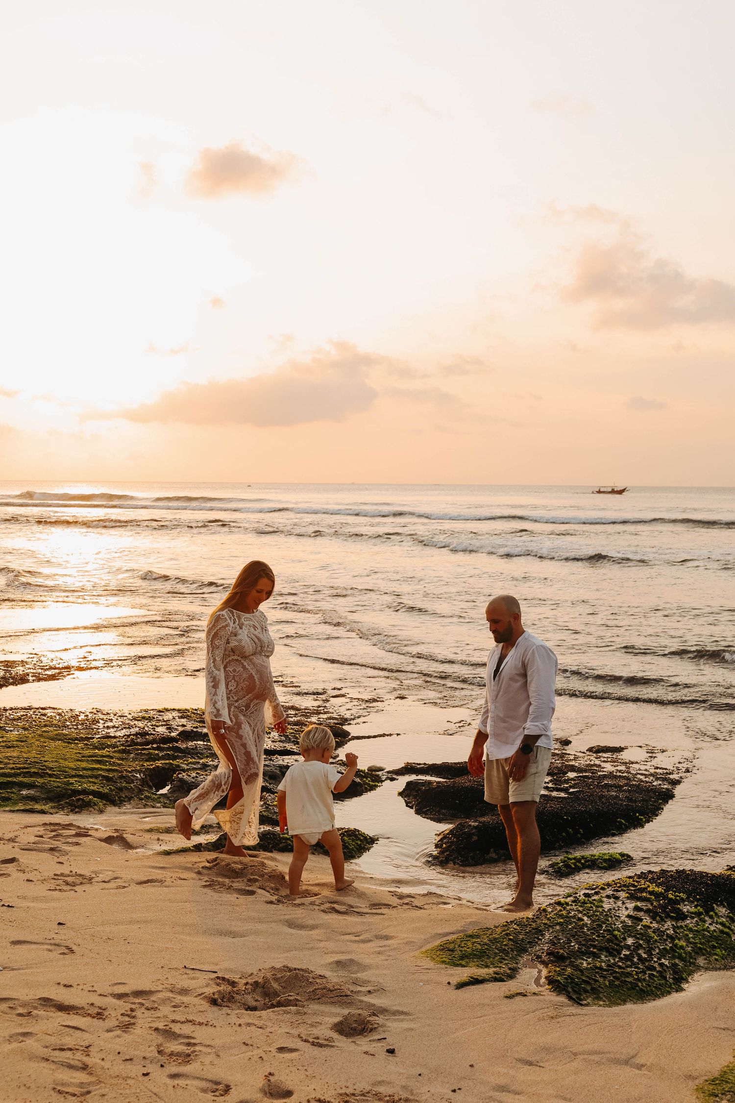 Pregnant woman and partner playing with toddler on sandy beach at sunset.