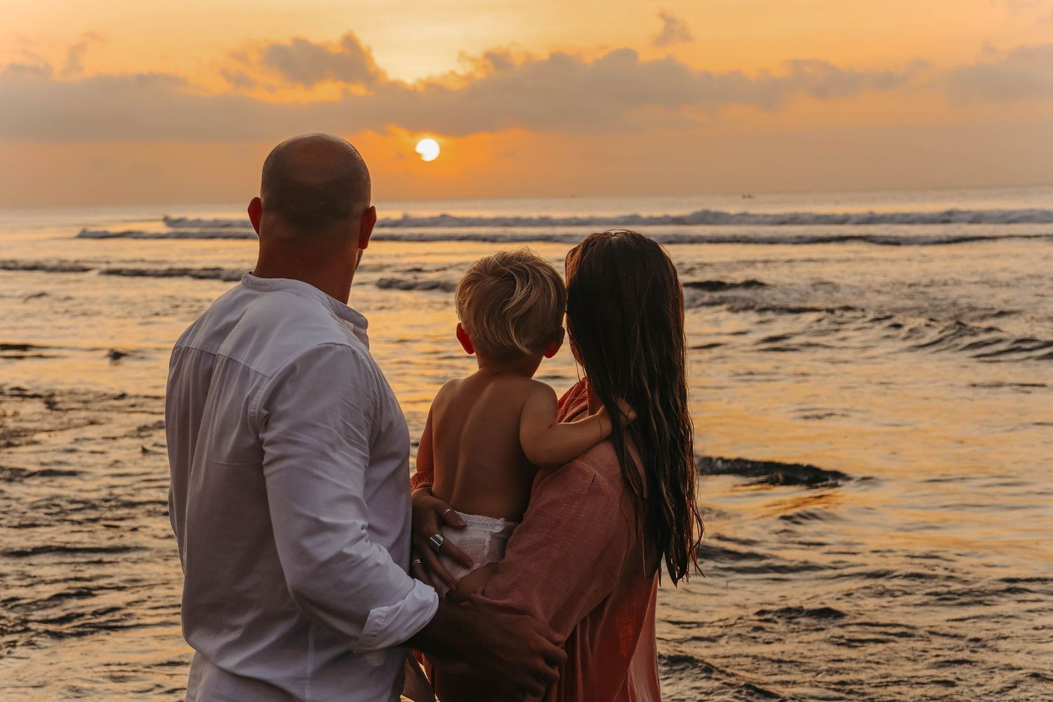 Family with child watching breathtaking orange sunset over ocean from beach.