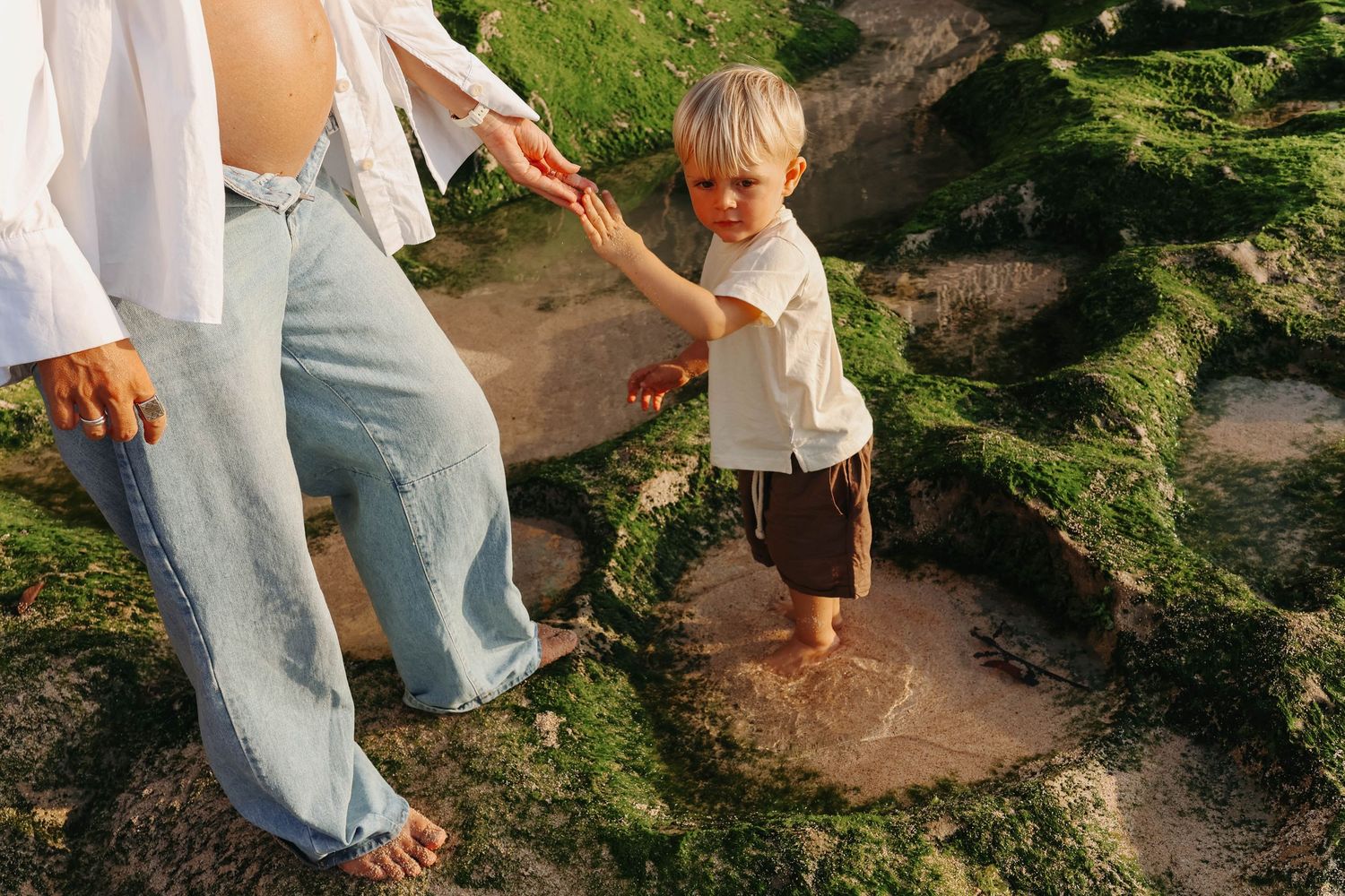 Pregnant mother helping toddler explore mossy rocks at beach during sunset.