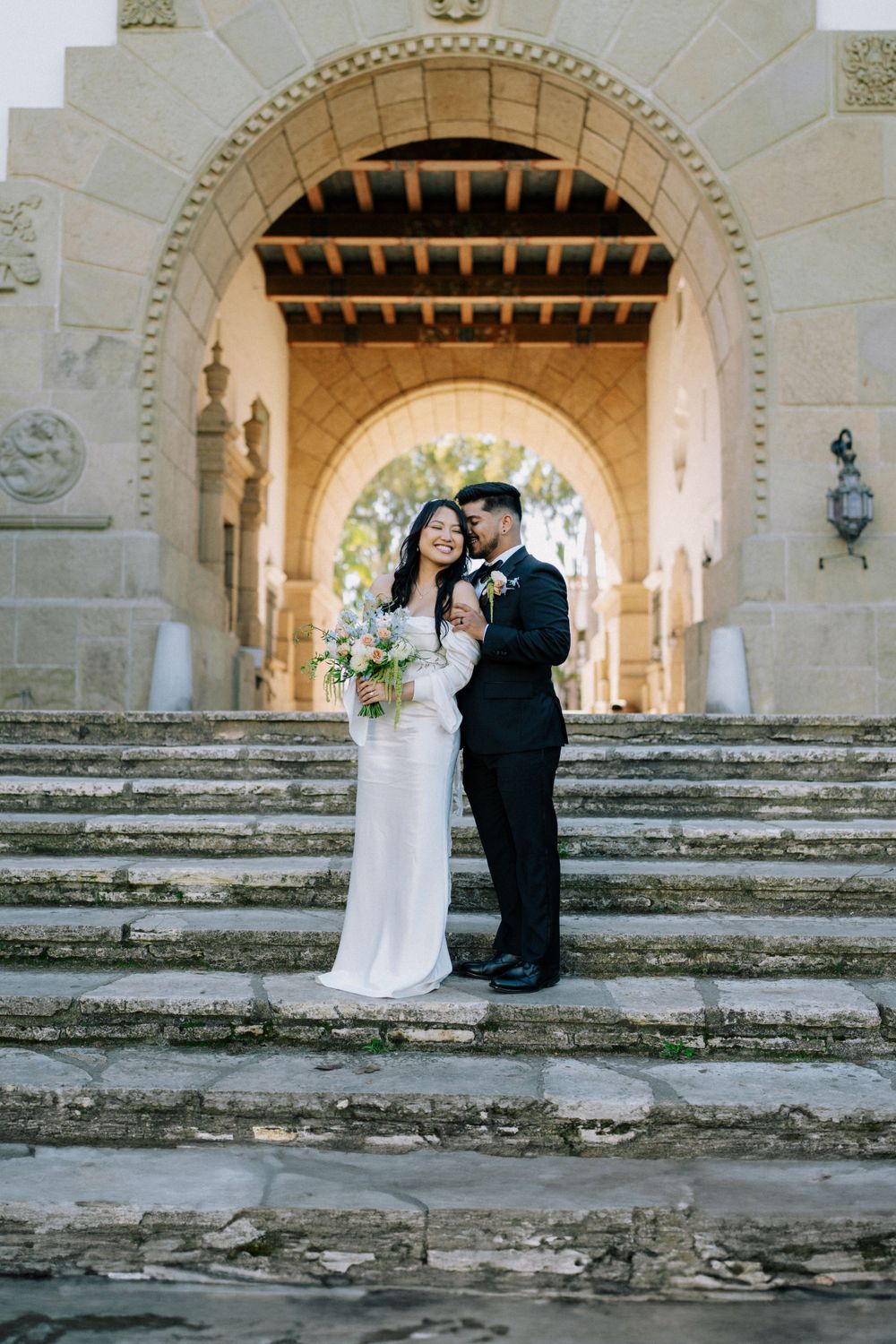 Elegant couple standing on stone steps beneath a Spanish-style archway.