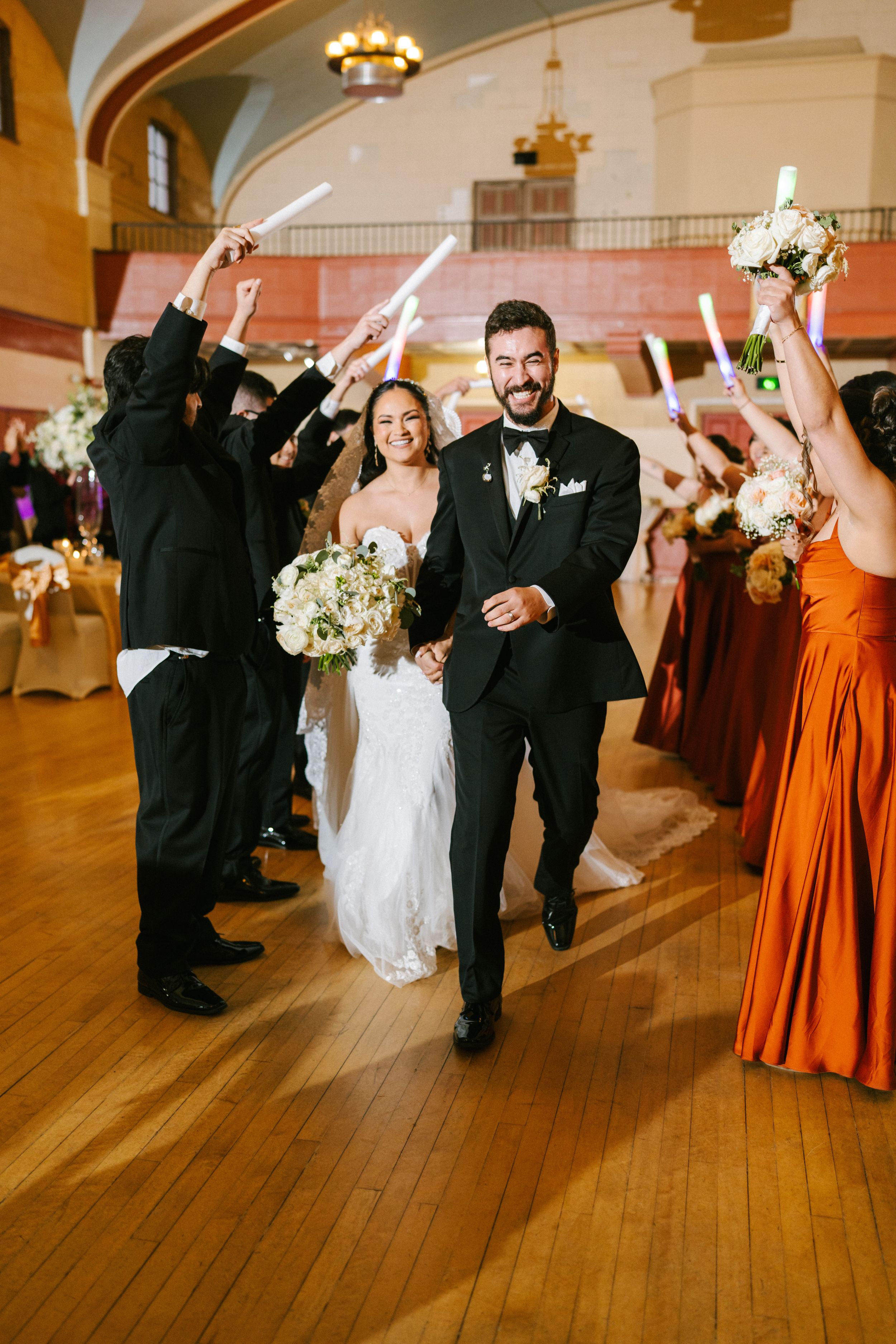 Wedding party dancing and celebrating on a wooden dance floor in a reception hall with orange bridesmaid dresses.