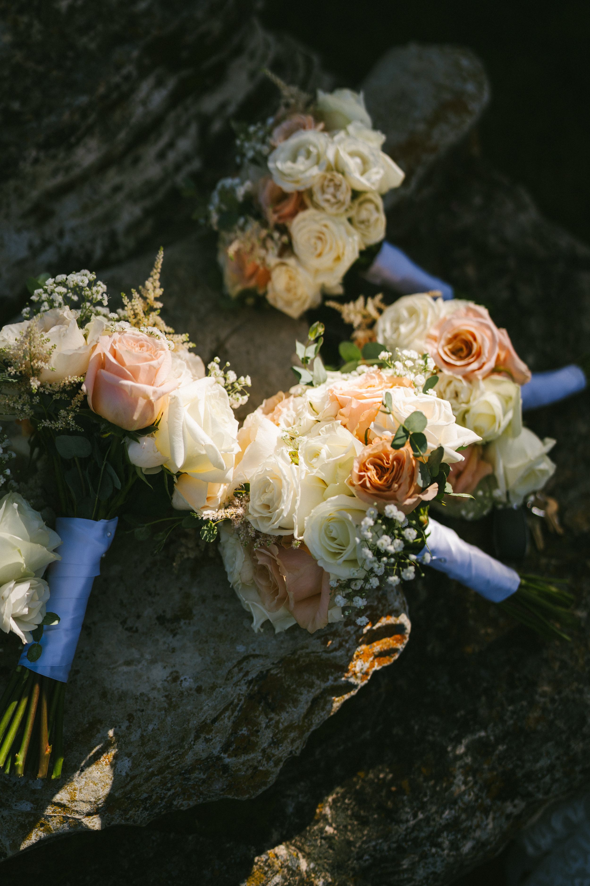 Wedding bouquets with peach and cream roses wrapped in blue ribbons rest on a dark stone surface in natural lighting.