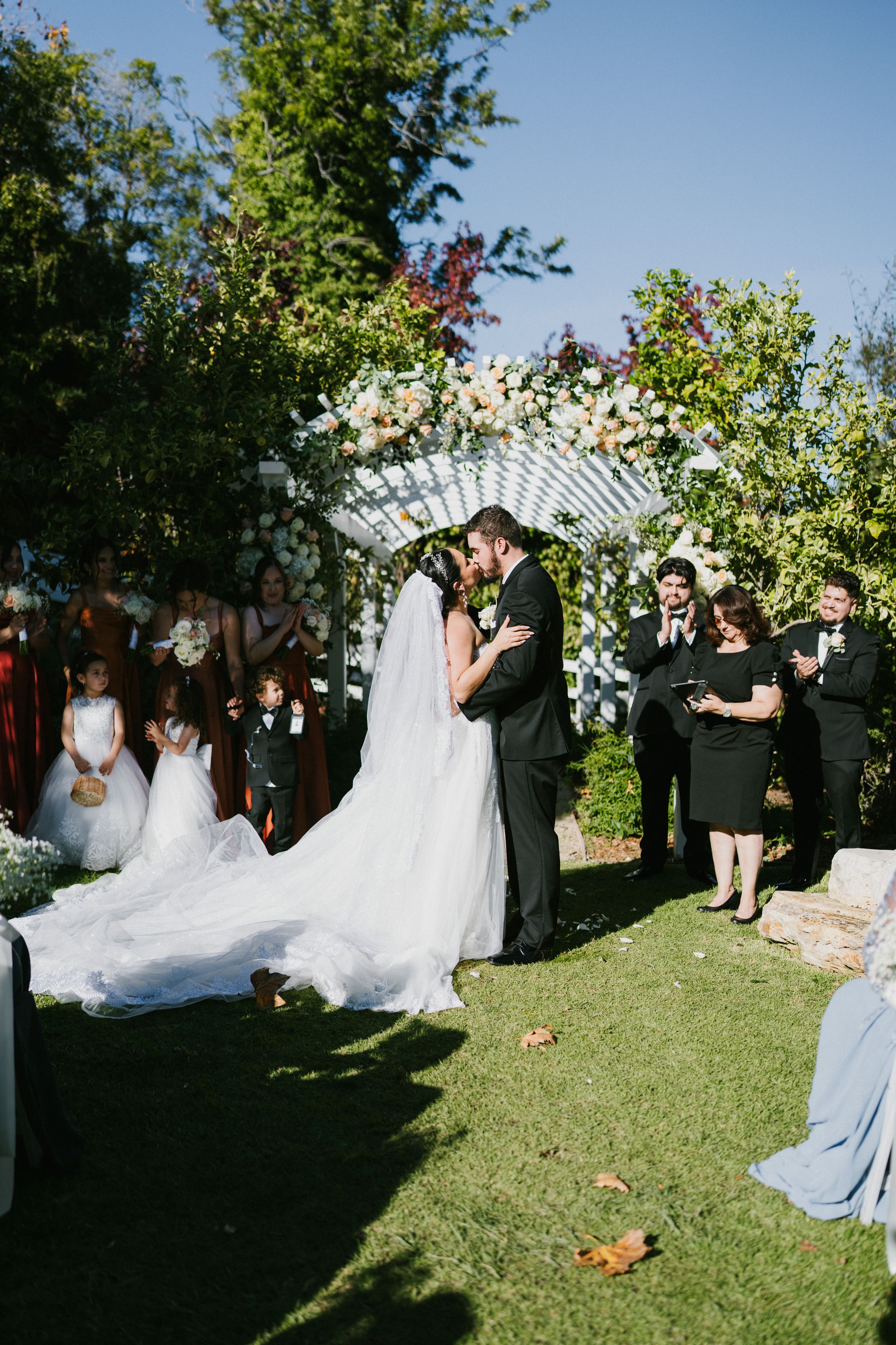 Wedding ceremony takes place outdoors under a white arch with guests gathered on a grassy lawn.