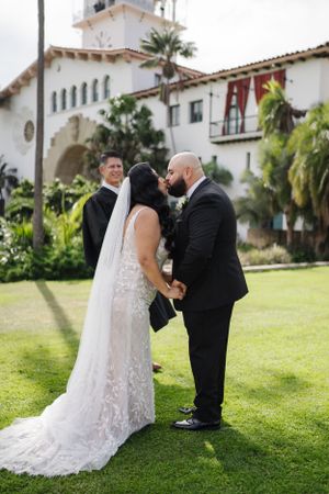 Wedding ceremony taking place on sunny lawn with Spanish-style building backdrop.