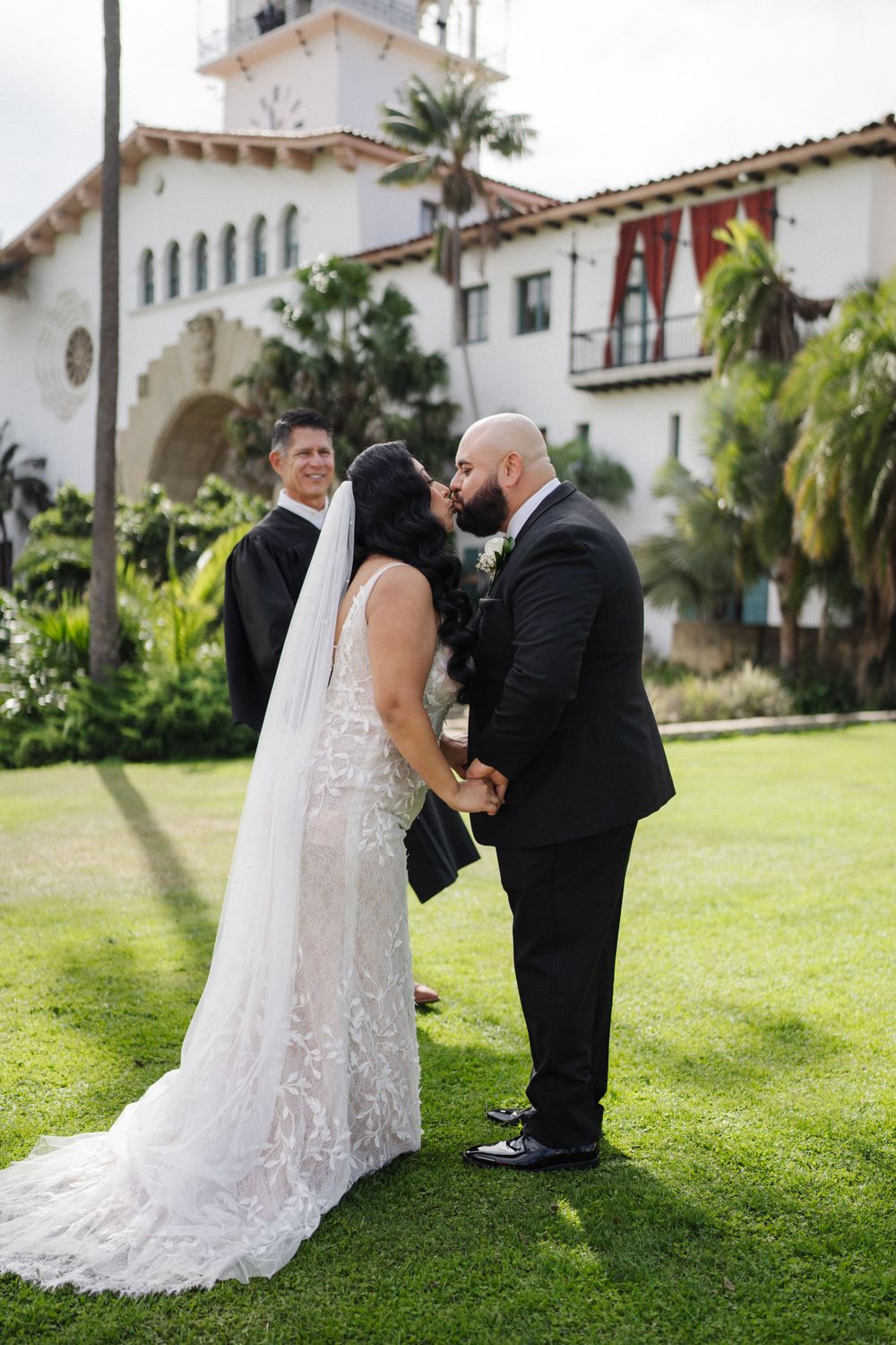 Wedding ceremony taking place on sunny lawn with Spanish-style building backdrop.