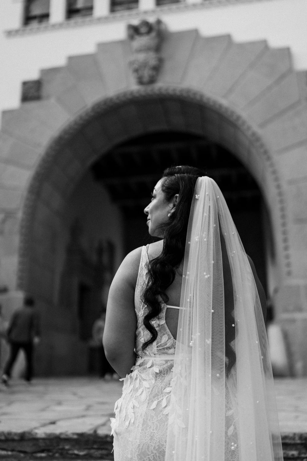 Black and white portrait of bride with flowing veil under historic archway.