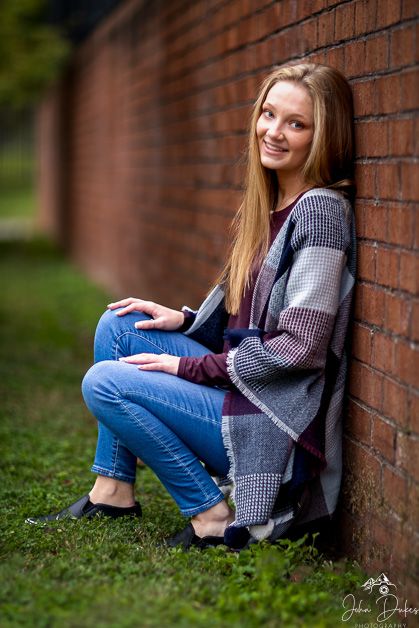 Senior portrait of person in plaid jacket and jeans sitting against brick wall.
