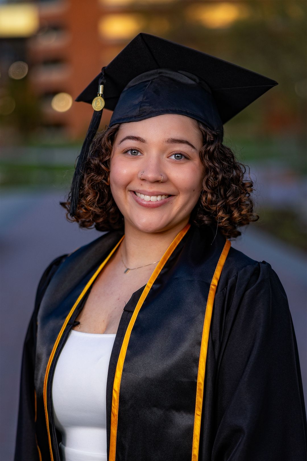 Smiling graduate in black cap and gown with gold stole poses outdoors on campus for graduation photo.