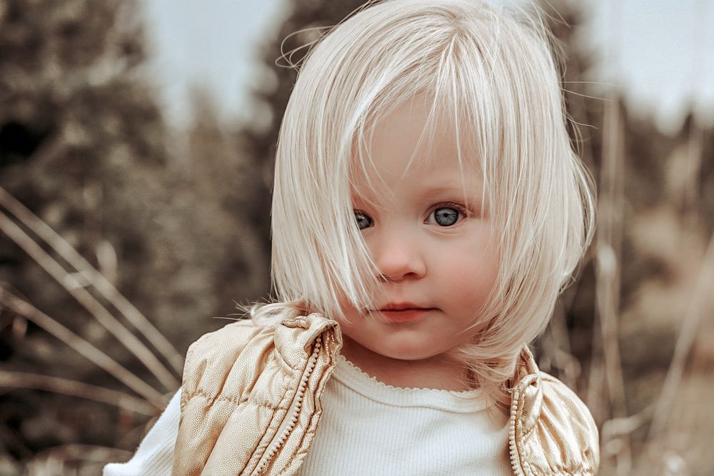 Portrait of a toddler with platinum blonde hair wearing a cream-colored outfit against a natural outdoor background.