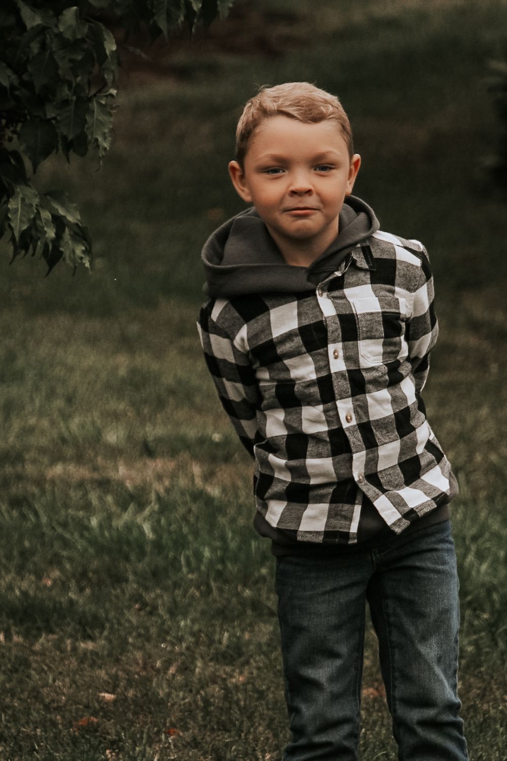 Young child wearing black and white buffalo plaid hoodie and jeans poses playfully outdoors.