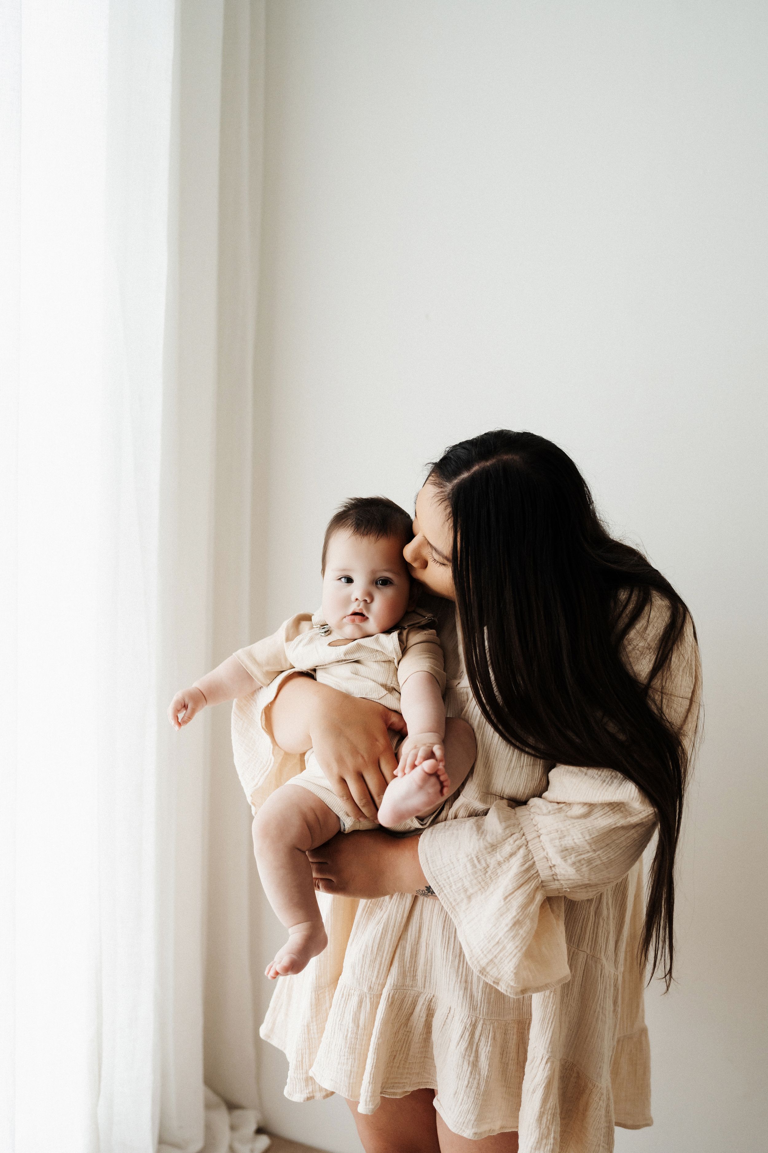 Gentle motherhood moments captured in soft natural light by white curtained windows.