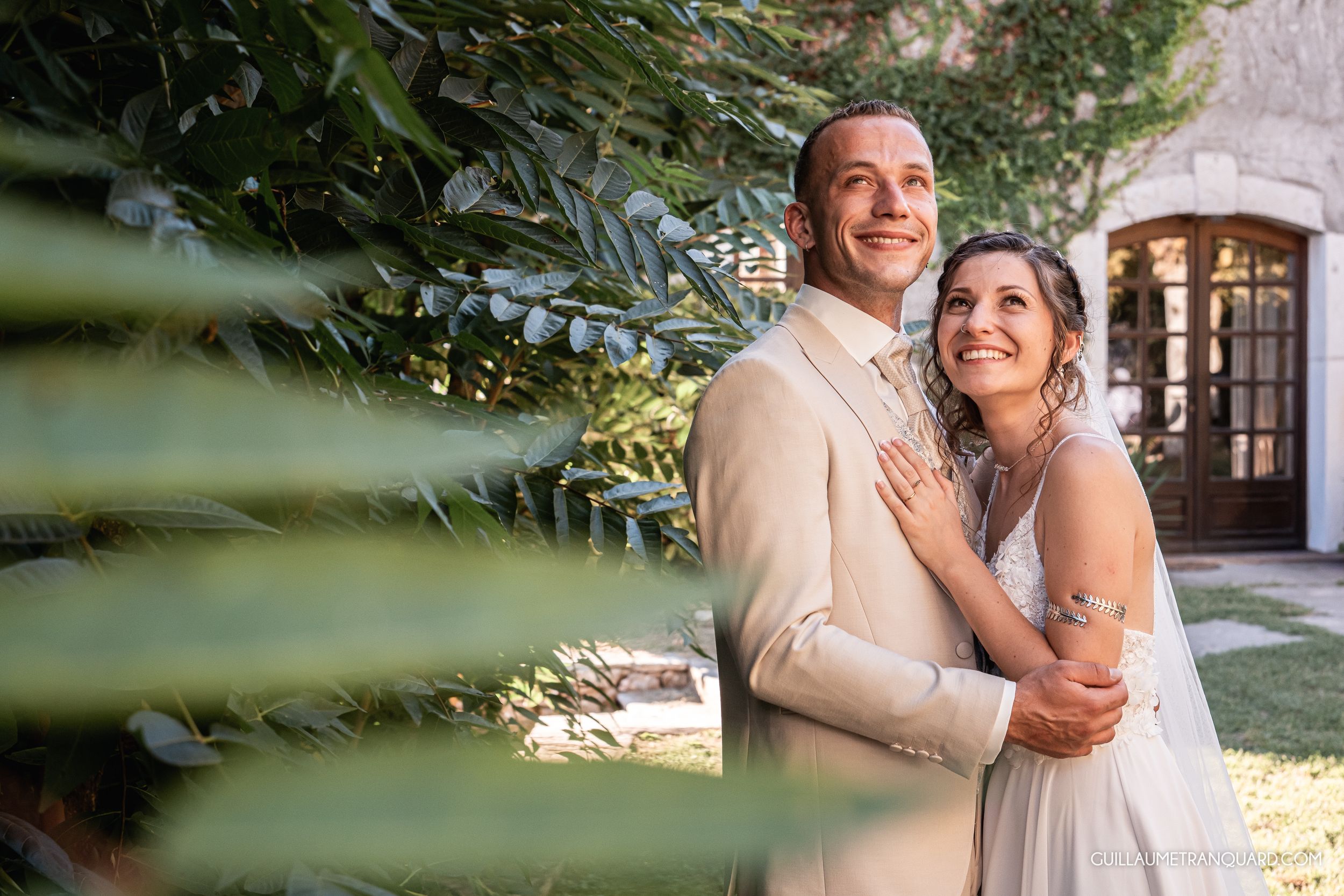 Smiling bride in a white gown embraces her groom in a beige suit in a sunlit garden framed by green tropical leaves.