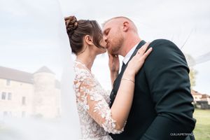 Bride in a lace wedding gown kisses her groom in a dark suit beneath a sheer veil, with a historic stone building behind them.