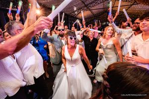 Bride in a white gown and sunglasses dances with joyful wedding guests who wave glow sticks on a lively reception dance floor.