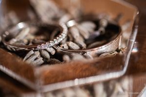 Two wedding rings, one with a sparkling diamond band, resting in a glass dish filled with dried seed pods.