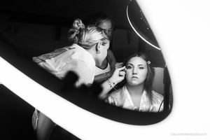 Black and white photo of a makeup artist applying makeup to a bride in a robe, reflected in a lit mirror during wedding preparations.