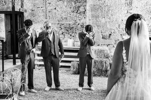 Three men in suits wipe away tears and cover their faces as they watch a bride in a lace gown and veil in a rustic stone venue.