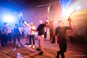 Guests wearing white masks hold lit sparklers and bottles while dancing on a wooden floor at a lively party in a stone-walled venue.