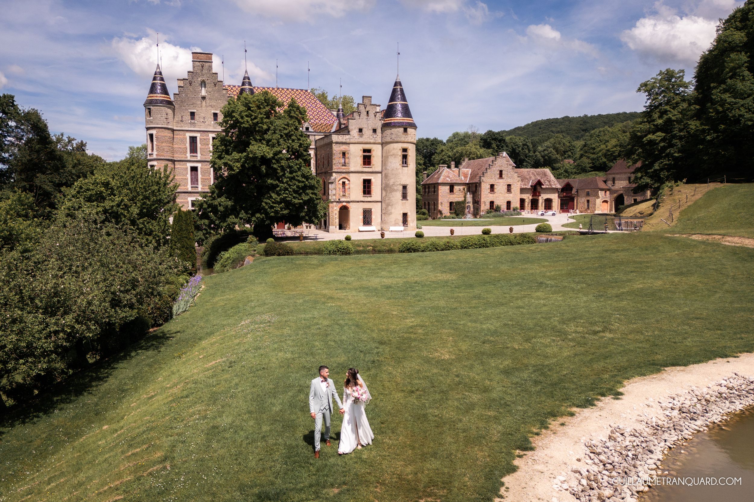 Couple in wedding attire walking on green lawn before a grand castle surrounded by lush trees on a sunny day.